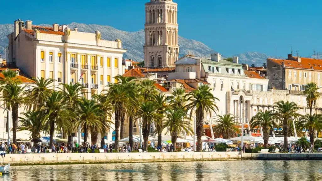 The Riva waterfront in Split seen from the harbour, with palm trees, the cathedral bell tower and the mountains behind