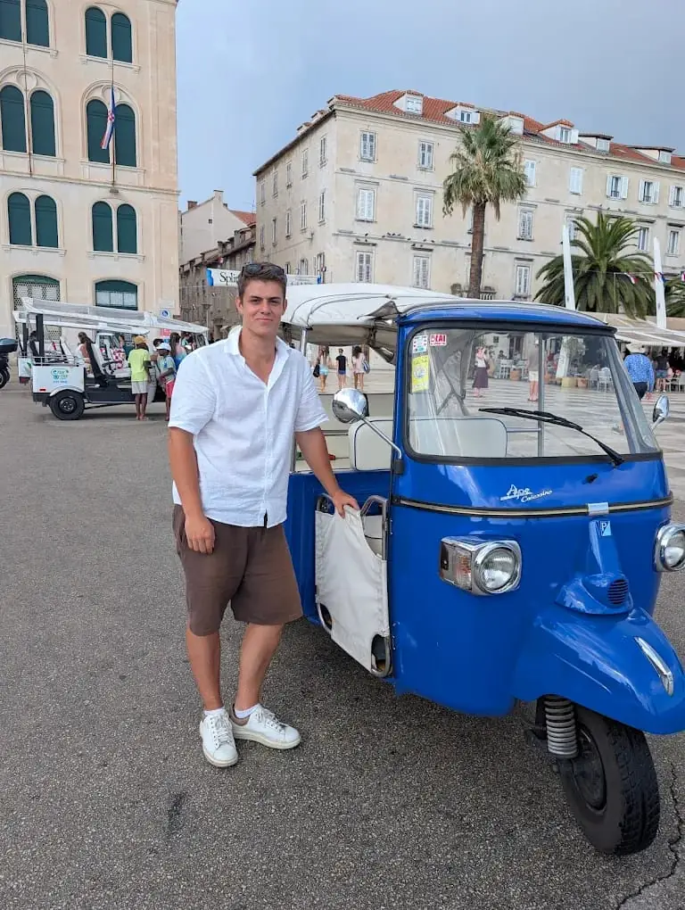 A guest posing next to a blue tuk tuk on Split's palm-lined Riva